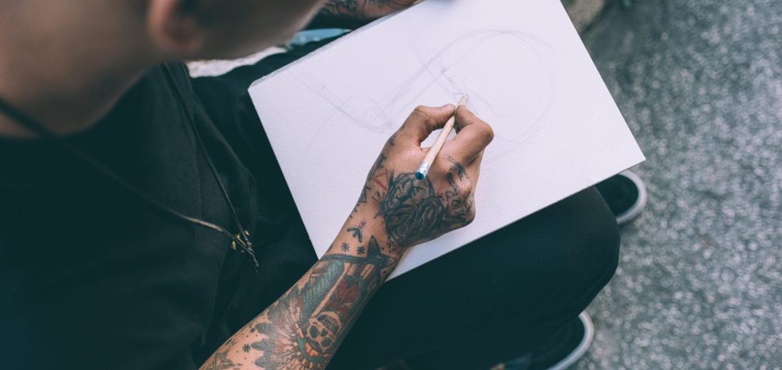 Close up on the hands of young hispanic man tattoed sketching on sketchbook - artist, drawning, creative concept